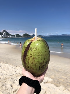 A hand is holding a green coconut with a straw inserted into it, against the backdrop of a sandy beach and clear blue sky. In the distance, a few people are visible near the ocean, some of them swimming. The shoreline has city buildings and mountains in the horizon.