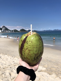 A hand is holding a green coconut with a straw inserted into it, against the backdrop of a sandy beach and clear blue sky. In the distance, a few people are visible near the ocean, some of them swimming. The shoreline has city buildings and mountains in the horizon.