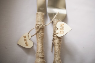 Two utensils wrapped in twine, with wooden heart tags attached. One tag reads '08-25-2018' and the other 'D&N'. The utensils are laid on a plain surface.
