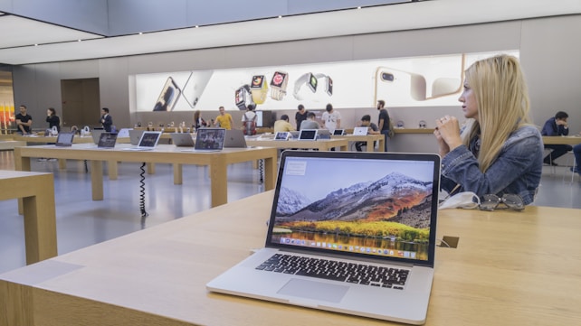 The image features a modern electronics store interior with wooden tables displaying various laptop models. Several people are present, some trying out devices while others converse. Large promotional images of smartwatches and smartphones are visible on the walls, adding to the store's sleek and innovative atmosphere.
