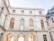 A large, elegant building features classic architectural design with arched windows and intricate carvings. The walls are light-colored stone, with a symmetrical layout and a series of large, golden-framed windows. The scene is bathed in soft daylight, creating a serene, historical atmosphere.