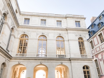 A large, elegant building features classic architectural design with arched windows and intricate carvings. The walls are light-colored stone, with a symmetrical layout and a series of large, golden-framed windows. The scene is bathed in soft daylight, creating a serene, historical atmosphere.