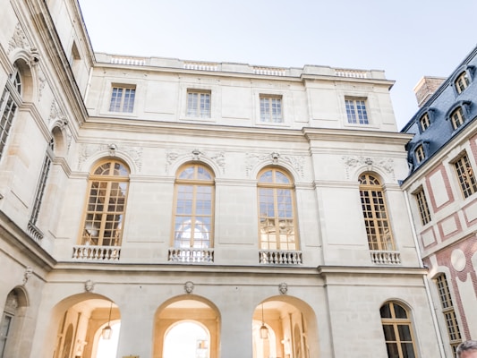 A large, elegant building features classic architectural design with arched windows and intricate carvings. The walls are light-colored stone, with a symmetrical layout and a series of large, golden-framed windows. The scene is bathed in soft daylight, creating a serene, historical atmosphere.