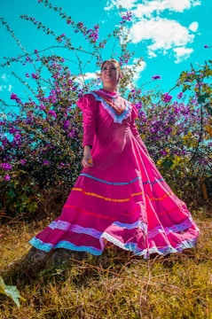 A woman stands confidently in a vibrant pink traditional dress with colorful embroidery and lace details. She is surrounded by lush purple flowers and green foliage under a bright blue sky with scattered clouds.