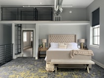 A modern bedroom features a large, tufted headboard with a beige upholstered bed. The bed is neatly made with white pillows and a beige throw. On either side, there are nightstands with lamps. Large windows with dark blinds allow natural light in. The room has a distinctive loft area with black railings accessed by a metal ladder. The flooring is a mix of dark gray and yellow patterned carpet.