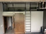 A modern interior space features a wooden sliding door with a metal track system, accompanied by a dark metal ladder leading to an overhead loft area. The loft is surrounded by metal railings and appears to contain a bed and some other furnishings. The floor outside the loft area appears to be tiled. Visible underneath the loft, a hallway extends into another part of the space. A lamp or chandelier with multiple bulbs is visible in the upper right corner.