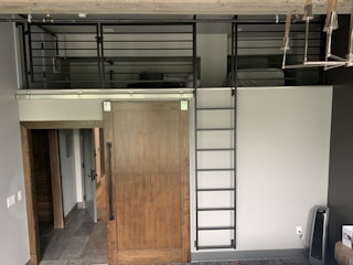 A modern interior space features a wooden sliding door with a metal track system, accompanied by a dark metal ladder leading to an overhead loft area. The loft is surrounded by metal railings and appears to contain a bed and some other furnishings. The floor outside the loft area appears to be tiled. Visible underneath the loft, a hallway extends into another part of the space. A lamp or chandelier with multiple bulbs is visible in the upper right corner.