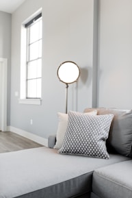 A modern living room with a minimalistic design features a light gray sectional sofa adorned with two decorative pillows, one in a textured pattern. A tall, circular floor lamp with a mid-century modern style stands beside the sofa. Natural light streams in through a large window, illuminating the wooden floors and pale gray walls, creating a serene and cozy atmosphere.