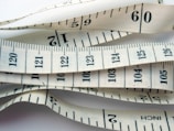 White measuring tape with black markings and numbers, displaying both inches and centimeters. It is coiled and layered, creating a visual pattern of overlapping tape displaying various numbers and measurement lines.