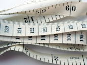 White measuring tape with black markings and numbers, displaying both inches and centimeters. It is coiled and layered, creating a visual pattern of overlapping tape displaying various numbers and measurement lines.
