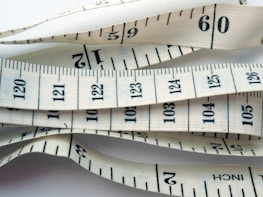 White measuring tape with black markings and numbers, displaying both inches and centimeters. It is coiled and layered, creating a visual pattern of overlapping tape displaying various numbers and measurement lines.