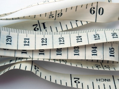 White measuring tape with black markings and numbers, displaying both inches and centimeters. It is coiled and layered, creating a visual pattern of overlapping tape displaying various numbers and measurement lines.