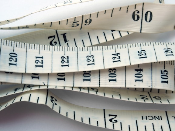 White measuring tape with black markings and numbers, displaying both inches and centimeters. It is coiled and layered, creating a visual pattern of overlapping tape displaying various numbers and measurement lines.