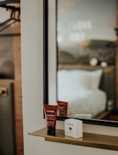 A sleek mirror with a minimalist shelf holds skincare products, including a tube labeled Lather moisturizer. The background displays a blurred view of a hotel room with a bed and a wall with indiscernible text.