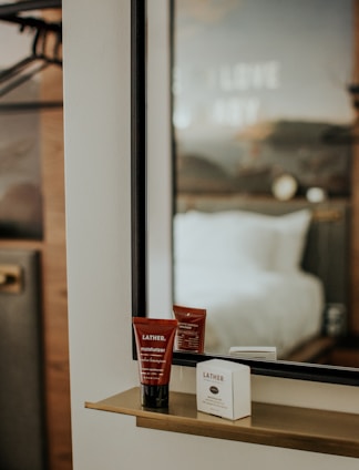 A sleek mirror with a minimalist shelf holds skincare products, including a tube labeled Lather moisturizer. The background displays a blurred view of a hotel room with a bed and a wall with indiscernible text.