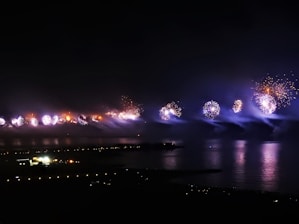 Fireworks illuminate the night sky over a body of water, creating vibrant reflections on the surface. The pyrotechnics display features bursts of light in various colors, set against a dark background with a distant view of city lights and buildings. Smoke trails from the fireworks add a misty effect.