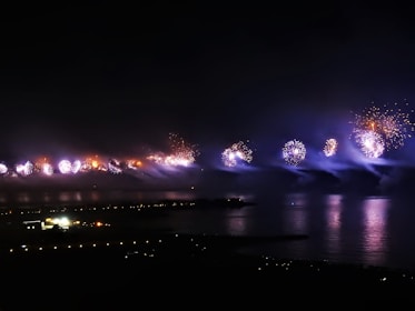 Fireworks illuminate the night sky over a body of water, creating vibrant reflections on the surface. The pyrotechnics display features bursts of light in various colors, set against a dark background with a distant view of city lights and buildings. Smoke trails from the fireworks add a misty effect.