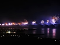 Fireworks illuminate the night sky over a body of water, creating vibrant reflections on the surface. The pyrotechnics display features bursts of light in various colors, set against a dark background with a distant view of city lights and buildings. Smoke trails from the fireworks add a misty effect.