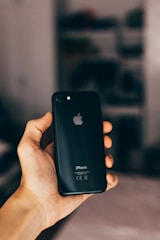 A hand holding a black smartphone with an Apple logo, set against a softly blurred background. The phone is an iPhone model.