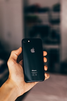 A hand holding a black smartphone with an Apple logo, set against a softly blurred background. The phone is an iPhone model.