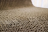 A close-up view of corrugated cardboard with a focus on its ridged texture. The light brown material creates a visually interesting pattern with its repetitive and undulating structure.