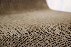 A close-up view of corrugated cardboard with a focus on its ridged texture. The light brown material creates a visually interesting pattern with its repetitive and undulating structure.