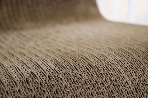 A close-up view of corrugated cardboard with a focus on its ridged texture. The light brown material creates a visually interesting pattern with its repetitive and undulating structure.