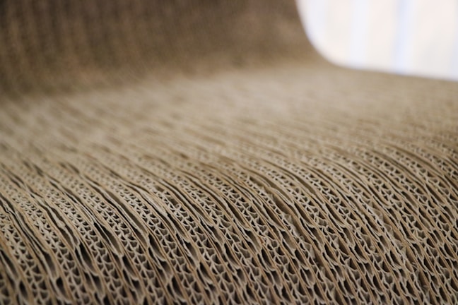 A close-up view of corrugated cardboard with a focus on its ridged texture. The light brown material creates a visually interesting pattern with its repetitive and undulating structure.