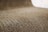 A close-up view of corrugated cardboard with a focus on its ridged texture. The light brown material creates a visually interesting pattern with its repetitive and undulating structure.