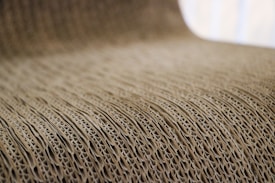 A close-up view of corrugated cardboard with a focus on its ridged texture. The light brown material creates a visually interesting pattern with its repetitive and undulating structure.