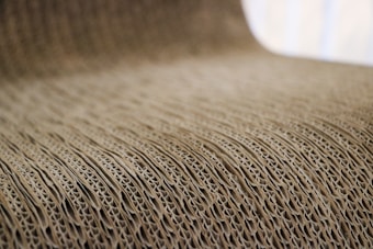 A close-up view of corrugated cardboard with a focus on its ridged texture. The light brown material creates a visually interesting pattern with its repetitive and undulating structure.