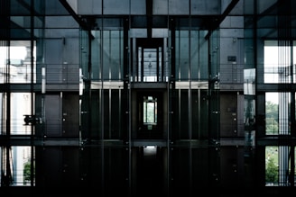 A modern architectural interior featuring large glass walls and metal structures. The space includes multiple levels with visible railings and corridors. The lighting is dim with natural light seeping in, creating reflective surfaces and shadows.