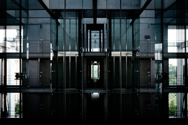 A modern architectural interior featuring large glass walls and metal structures. The space includes multiple levels with visible railings and corridors. The lighting is dim with natural light seeping in, creating reflective surfaces and shadows.
