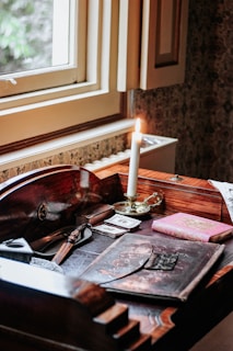 A wooden desk near a window covered with various vintage items including a lit candle in a brass holder, an ornate letter opener, old photographs, and a pink book with a decorative cover. The natural light from the window adds a warm atmosphere to the space.