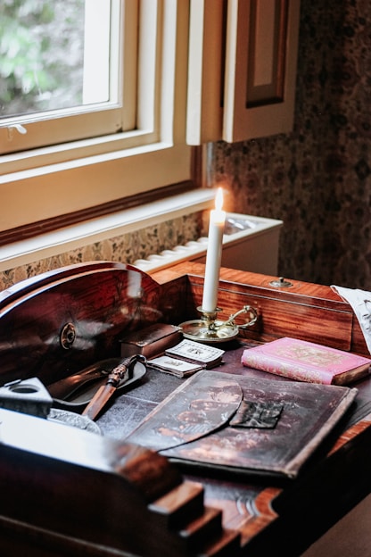A softly lit vintage wooden desk displaying an open handcrafted book with botanical illustrations, surrounded by delicate paper objects and a brass reading lamp.