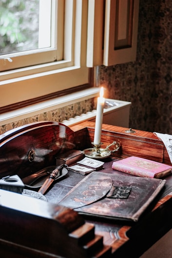 A wooden desk near a window covered with various vintage items including a lit candle in a brass holder, an ornate letter opener, old photographs, and a pink book with a decorative cover. The natural light from the window adds a warm atmosphere to the space.
