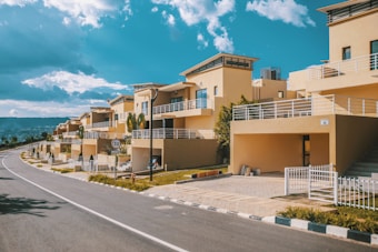 A row of modern, beige-colored two-story houses with flat roofs, large balconies, and fenced yards is situated along a curved, empty roadway under a partly cloudy blue sky. The area is landscaped with small trees and shrubs, and a speed limit sign is visible.