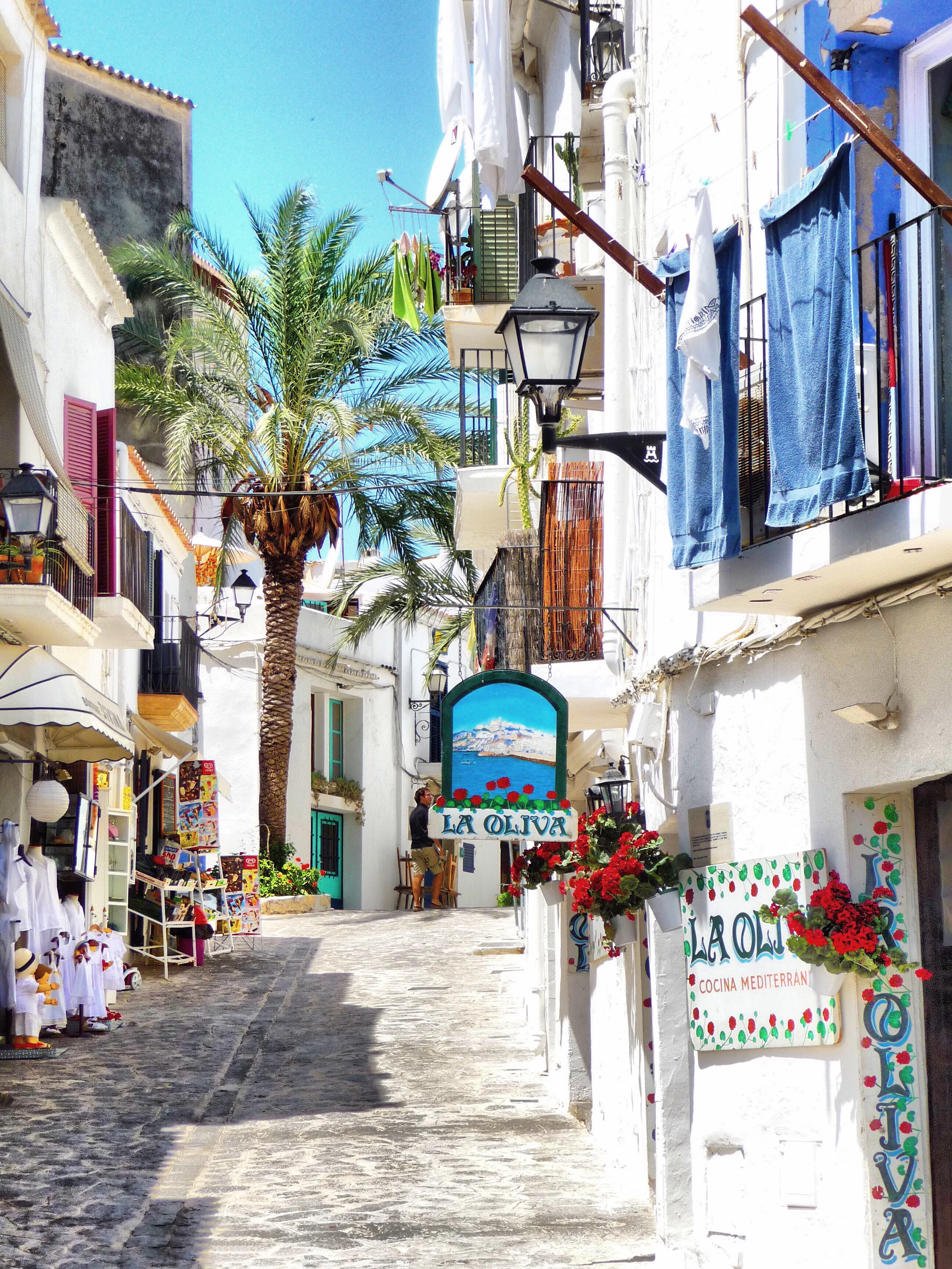 A narrow sunlit lane in Ibiza's old town with a palm tree, laundry and blue textiles hanging from balconies, and the hand-painted sign of the La Oliva restaurant overhead