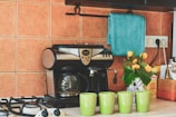 A cozy kitchen corner features a sleek, black and silver coffee maker beside four lime green mugs. On the counter, there is a decorative vase with bright yellow flowers, adding a touch of freshness. A teal towel hangs above, contrasting with the warm orange tiles of the backsplash. A box of consumables and a package is neatly arranged alongside the setup.