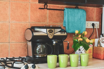 A cozy kitchen corner features a sleek, black and silver coffee maker beside four lime green mugs. On the counter, there is a decorative vase with bright yellow flowers, adding a touch of freshness. A teal towel hangs above, contrasting with the warm orange tiles of the backsplash. A box of consumables and a package is neatly arranged alongside the setup.