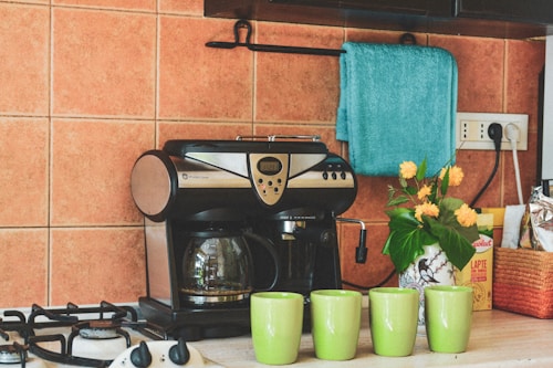 A cozy kitchen corner features a sleek, black and silver coffee maker beside four lime green mugs. On the counter, there is a decorative vase with bright yellow flowers, adding a touch of freshness. A teal towel hangs above, contrasting with the warm orange tiles of the backsplash. A box of consumables and a package is neatly arranged alongside the setup.