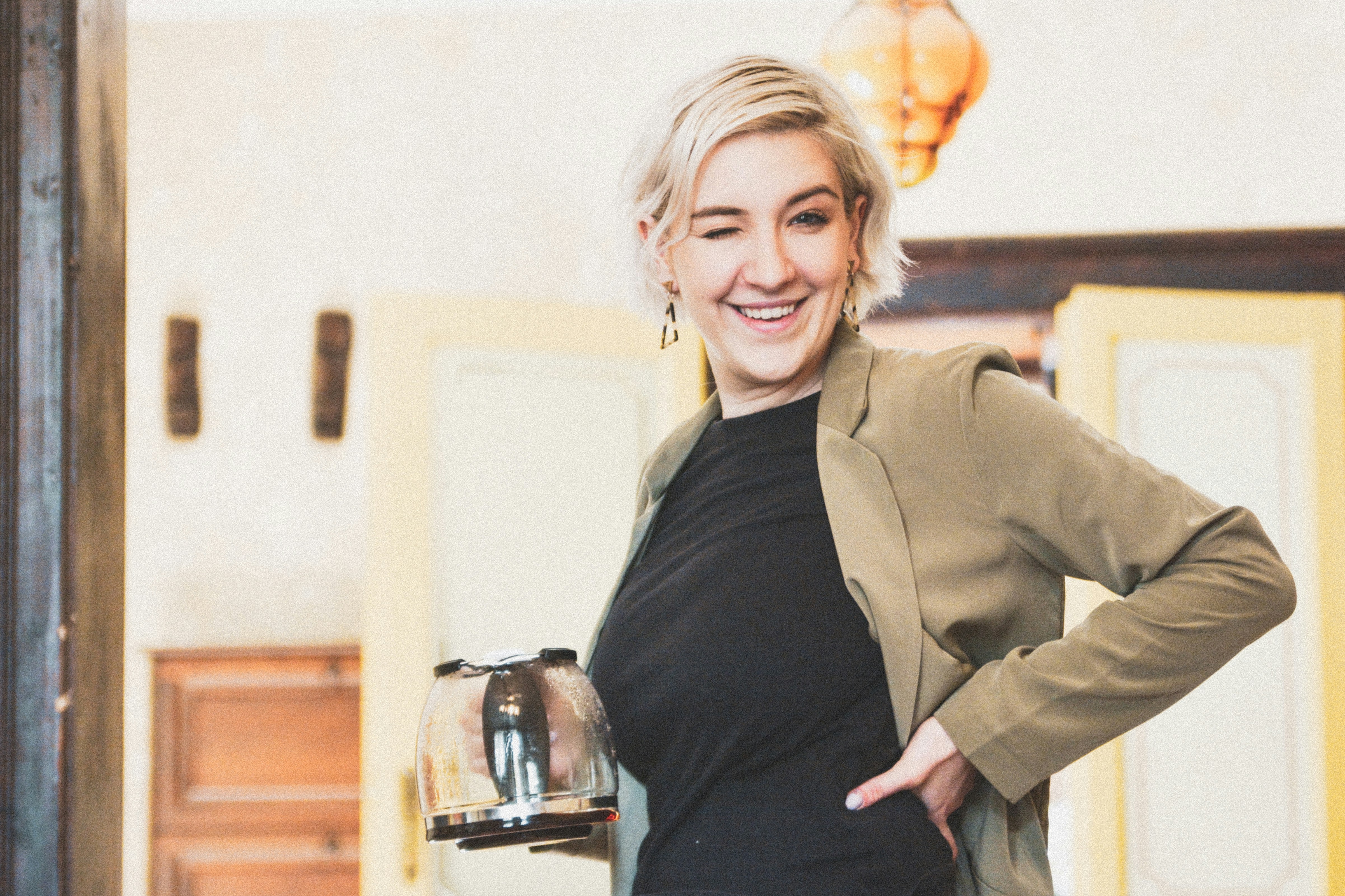 Lächelnde Frau in schwarzem Hemd und brauner Jacke, die mit dem linken Arm Akimbo steht