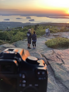 A couple is standing on a rocky hilltop, holding hands and facing a scenic view of the ocean with several small islands visible. The sun is setting on the horizon, casting a warm glow over the landscape. To the side, another couple is walking down the hill. A camera is prominently visible in the foreground, capturing the scene.
