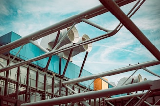 A modern architectural structure with large metal pipes and vents against a cloudy blue sky. The building features a combination of glass and metal, with prominent geometric shapes and a network of white beams crossing the composition.