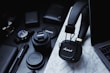 A collection of tech gadgets and accessories are arranged on a marble surface, including Marshall headphones, Canon camera lenses, a wristwatch, pens, and a laptop. The items are predominantly black and organized neatly, conveying a sense of modernity and style.