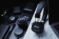 A collection of tech gadgets and accessories are arranged on a marble surface, including Marshall headphones, Canon camera lenses, a wristwatch, pens, and a laptop. The items are predominantly black and organized neatly, conveying a sense of modernity and style.