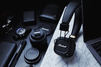 A collection of tech gadgets and accessories are arranged on a marble surface, including Marshall headphones, Canon camera lenses, a wristwatch, pens, and a laptop. The items are predominantly black and organized neatly, conveying a sense of modernity and style.