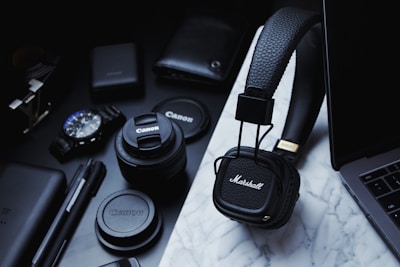 A collection of tech gadgets and accessories are arranged on a marble surface, including Marshall headphones, Canon camera lenses, a wristwatch, pens, and a laptop. The items are predominantly black and organized neatly, conveying a sense of modernity and style.