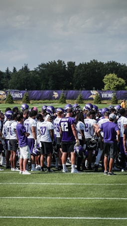 A group of football players huddled together on a grassy field, wearing jerseys and helmets. The players appear to be in a team meeting or discussion. Trees and a fencing banner with a team's name are visible in the background under a partly cloudy sky.