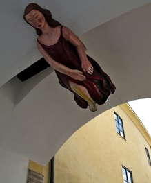 A ceiling-mounted figure of a woman in a flowing dark dress seems to be floating or suspended within an arched passageway. Below, part of a yellow building with several windows is visible. The name of the street, 'VIA PORTA SABIONA,' is displayed on a placard attached to the building.